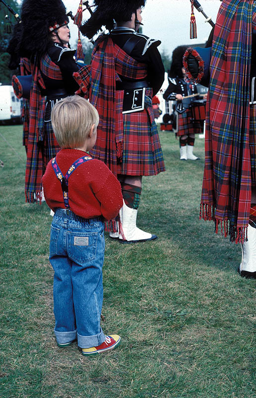 little boy standing behind some people in Scottish attire playing the bagpipes